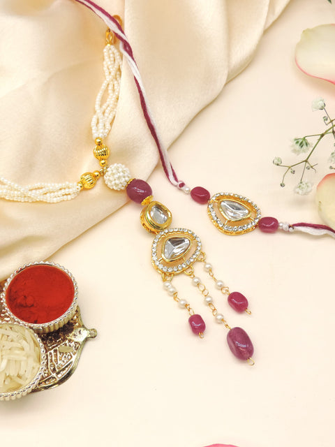 Gold and white beaded Rakhi with maroon beads and clear rhinestone heart-shaped details, displayed on a cream satin fabric alongside a silver-toned ring and decorative petals.