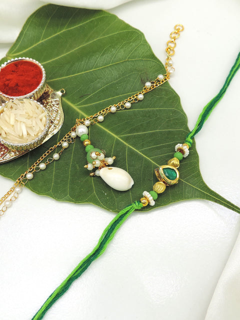 A traditional Indian Rakhi with a green cord and gold-toned charm, displayed on a large green leaf alongside a silver decorative plate with red powder and white rice on a white fabric background.