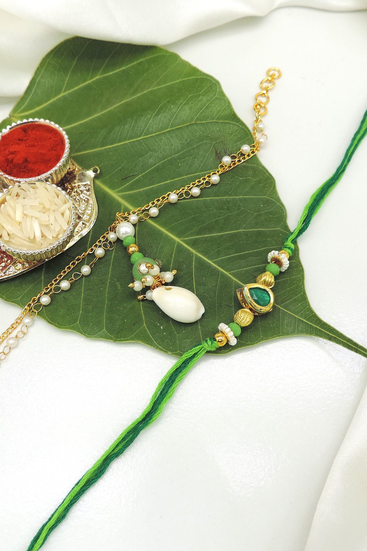 A traditional Indian Rakhi with a green cord and gold-toned charm, displayed on a large green leaf alongside a silver decorative plate with red powder and white rice on a white fabric background.